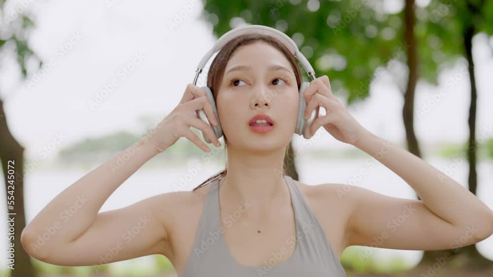Asian woman is engaged in fitness in public park. Female wearing sport bra and using headphone listening to music. Healthy lifestyle concept, sports outdoor activities in park