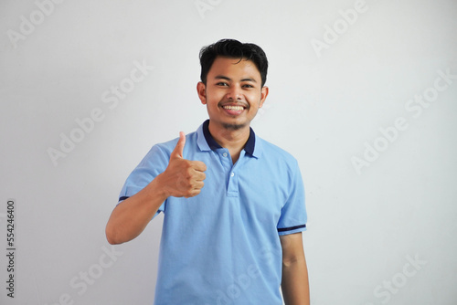 Portrait of cheerful asian man in wearing blue t shirt smiling and showing thumbs up at camera isolated over white background