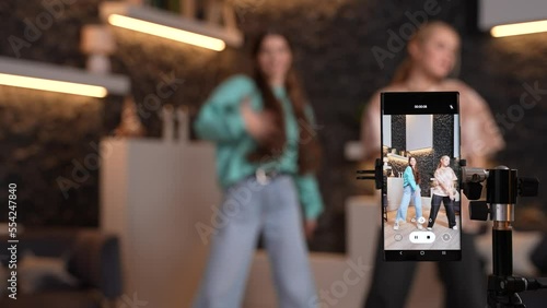 Female teens dancing together at home, recording a dance video for social media on smartphone camera. Young bloggers blurry on background making content for their channel. High quality footage.