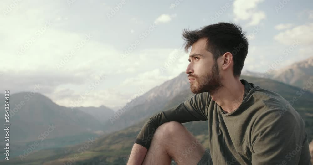 Portrait of an ethnic Caucasian man with a happy inspired bearded face. a young adult thoughtful guy is sitting on the top of a mountain against the background of the setting sun dreaming thinking Portrait of an ethnic Caucasian man with a happy inspired bearded face. a young adult thoughtful guy is sitting on the top of a mountain against the background of the setting sun dreaming thinking