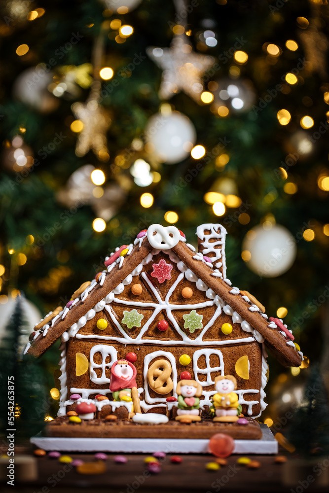 Christmas house made from gingerbread. Playfully decorated with colourful gummy bears. Christmas tree in background with lights. Festive New year atmosphere and mood. 