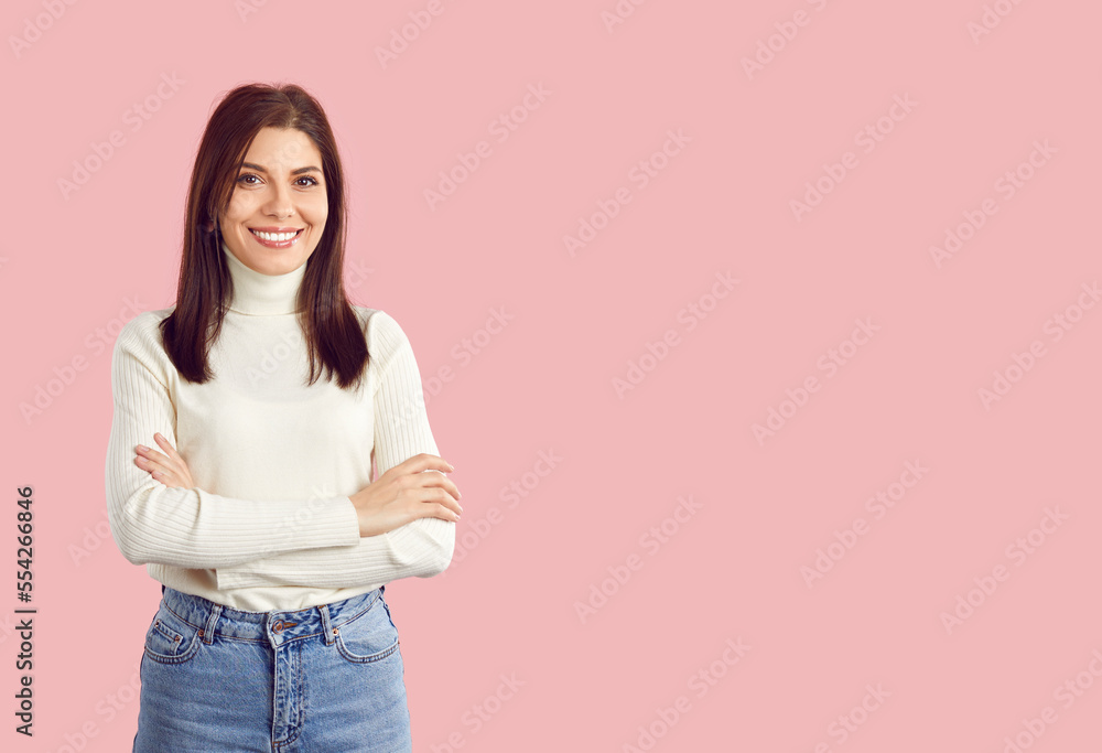 Portrait of happy beautiful smiling charming young brunette woman in casual white turtleneck and blue jeans standing with her arms crossed on empty solid light pink copy space studio background Portrait of happy beautiful smiling charming young brunette woman in casual white turtleneck and blue jeans standing with her arms crossed on empty solid light pink copy space studio background