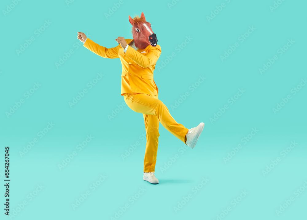 Stylish man in a funny horse mask and a bright yellow suit is fooling around, raising his hands up. Full-size photo of a funny guy dancing on an isolated turquoise background.