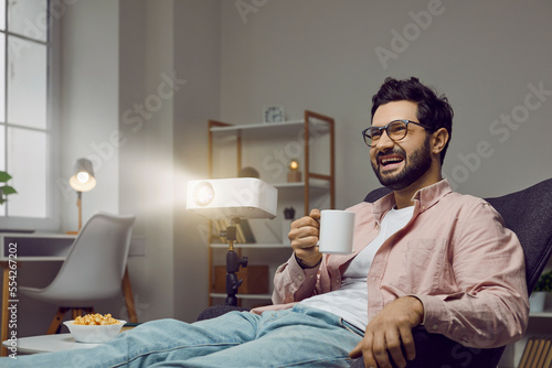 Evening film watch on projector at home. Cheerful man is watching comedy movie alone at home with help of movie projector. Portrait of young man sitting in chair with cup of hot drink and laughing.