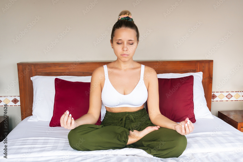 Young mindful attractive woman meditates holding fingers in yoga sign with her eyes closed sitting on white bed in bedroom. Peaceful girl keeps hands in mudra gesture sitting in the lotus position Young mindful attractive woman meditates holding fingers in yoga sign with her eyes closed sitting on white bed in bedroom. Peaceful girl keeps hands in mudra gesture sitting in the lotus position