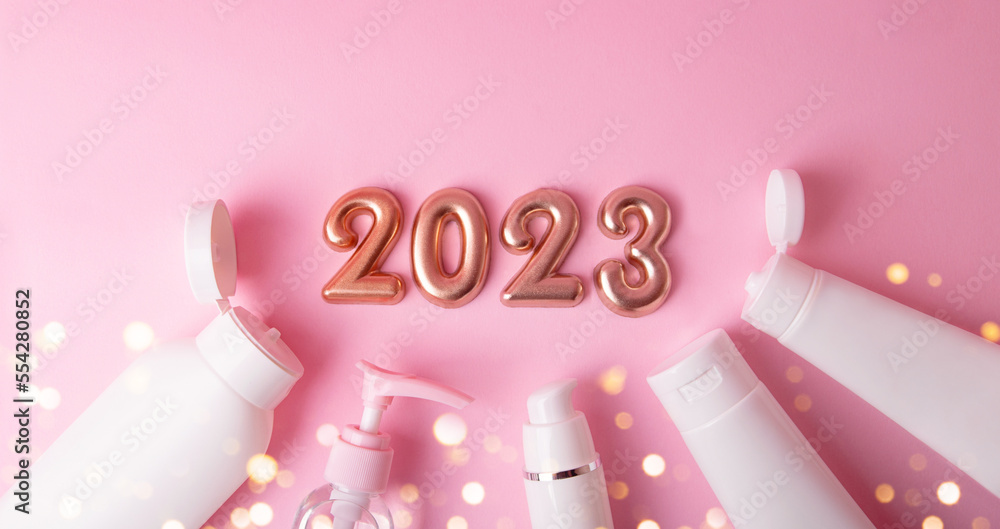 Top view of the cosmetics containers on pink background.Rose gold numbers 2023 above with shiny bokeh.