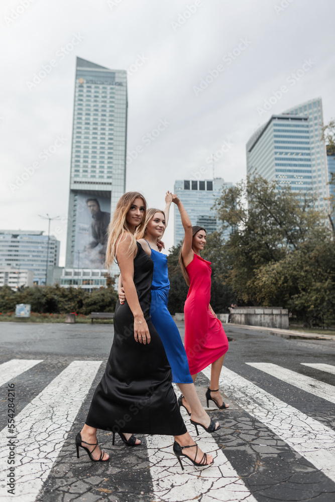 Beautiful fashionable girl friends in elegant clothes with dresses go to the crosswalk in the city