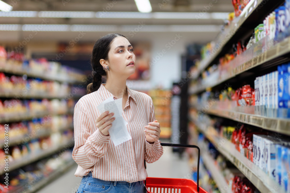 Portrait of young pretty caucasian woman holds checklist and choosing food and products. Showcase and shelves at background. Concept of shopping and consumerism