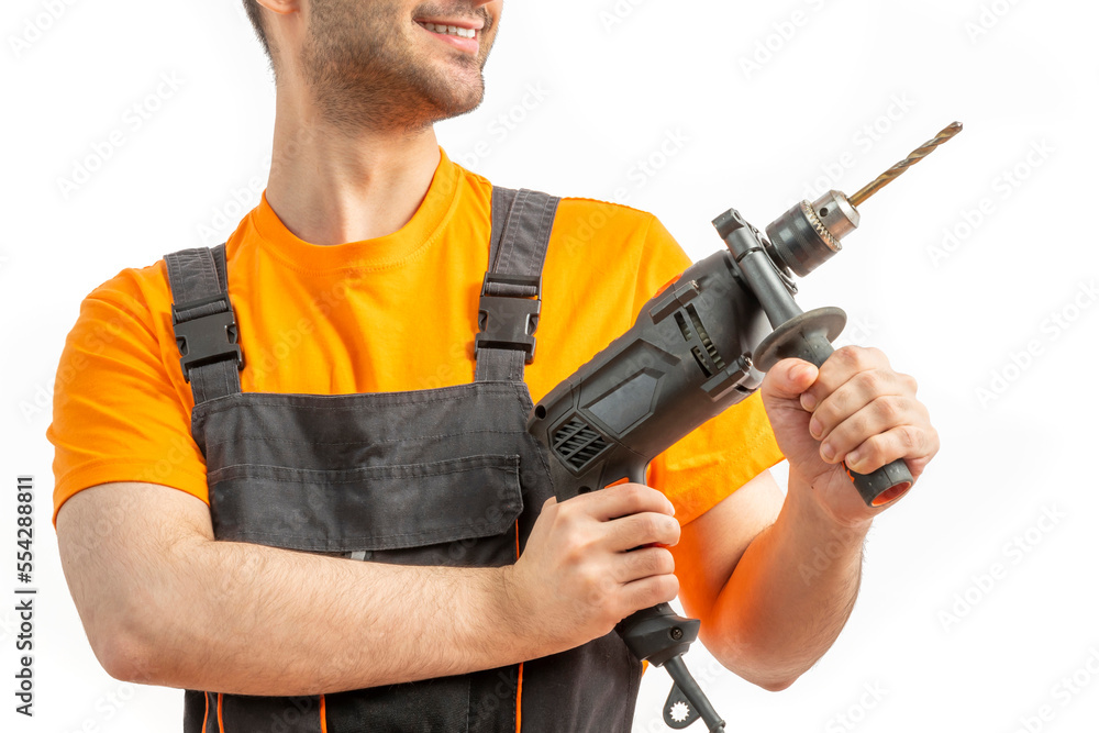 Young worker drilling with electric drill in his hands. Tool for boring holes. Craftsman repairman wearing orange uniform cap and t-shirt, overalls on a white isolated background for advertising Young worker drilling with electric drill in his hands. Tool for boring holes. Craftsman repairman wearing orange uniform cap and t-shirt, overalls on a white isolated background for advertising