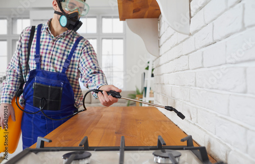 Pest control worker treating the inside of the house. Young man in a mask and blue workwear uniform working in a modern apartment, standing in the kitchen and spraying insecticide over the countertop