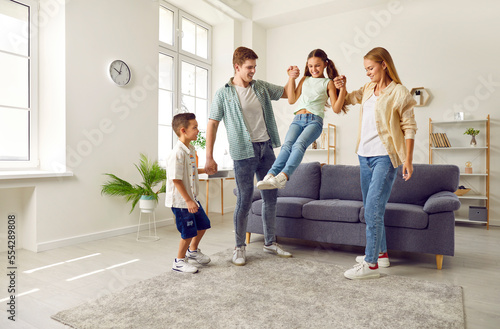 Happy family with two children are playing together. Girl is jumping from sofa holding hands her parents at home in living room. Mom, dad, son and daughter. Family lifestyle, love fun care concept.