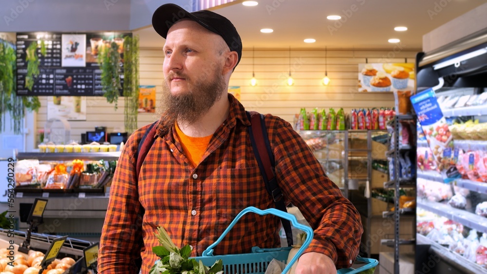 Bearded man in supermarket stands by shelves with groceries in his hand, basket of vegetables. Grocery shopping by millennial men in their 30s and 40s. A man chooses products in the store for cooking.