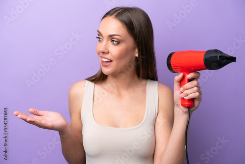 Young caucasian woman holding a hairdryer isolated on purple background with surprise facial expression