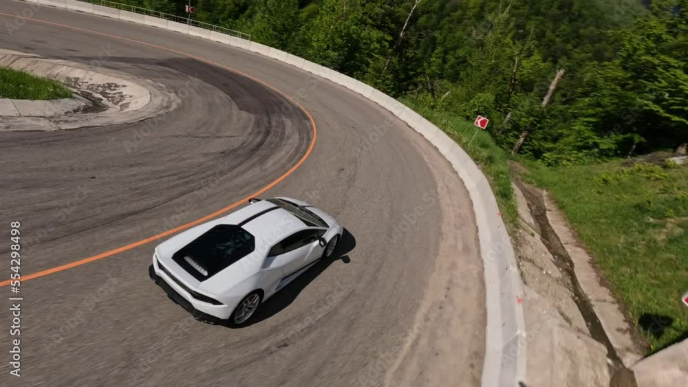 FPV sports drone white sport automobile riding on serpentine asphalt road mountain slope snowy peak sky landscape. Amazing summer alpine resort transport infrastructure architecture exterior at valley