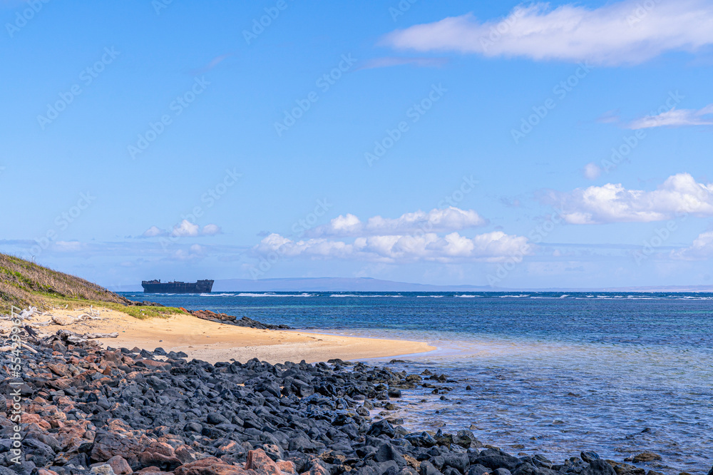 The rocky and sandy shore of Shipwreck Beach in Lanai with an old naval shipwreck grounded in the reef and the Island of Maui in the distance; Lanai, Hawaii, United States of America The rocky and sandy shore of Shipwreck Beach in Lanai with an old naval shipwreck grounded in the reef and the Island of Maui in the distance; Lanai, Hawaii, United States of America