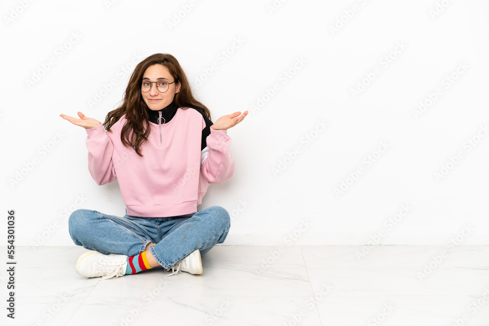 Young caucasian woman sitting on the floor isolated on white background having doubts while raising hands Young caucasian woman sitting on the floor isolated on white background having doubts while raising hands