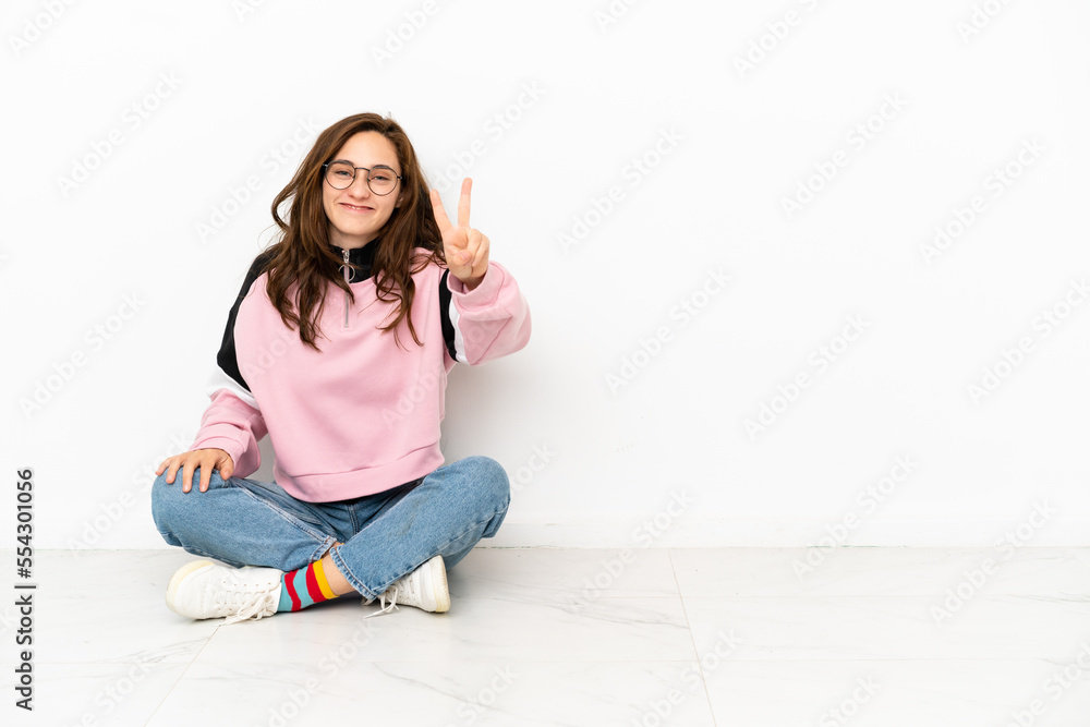 Young caucasian woman sitting on the floor isolated on white background smiling and showing victory sign