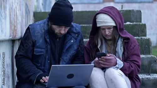 Two frozen homeless people, a man and a woman, sit in the street. They are holding a laptop and a mobile phone. The couple are looking for job advertisements.