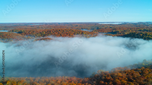 An aerial drone shot of fog coming off the lakes during the morning while a beautiful sunny Autumn Forest surrounds it