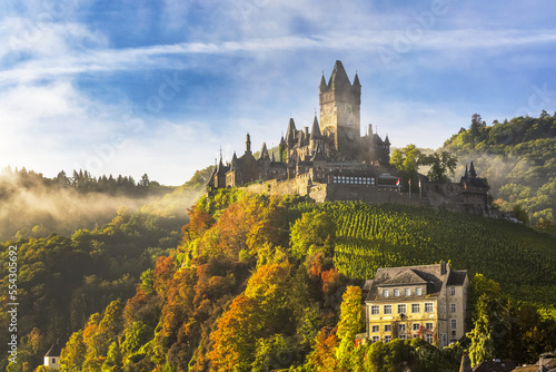 Large medieval castle on a colourful treed hillside with fog, blue sky and cloud; Cochem, Germany photo