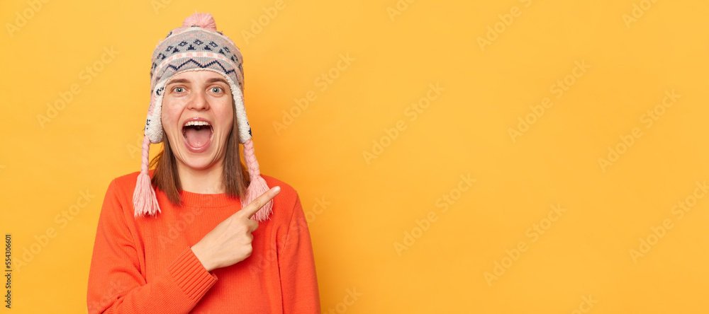 Portrait of amazed woman wearing orange jumper and knitted earflap hat standing isolated over yellow background, pointing away, showing free space, keeps mouth open, space for promotion. Portrait of amazed woman wearing orange jumper and knitted earflap hat standing isolated over yellow background, pointing away, showing free space, keeps mouth open, space for promotion.