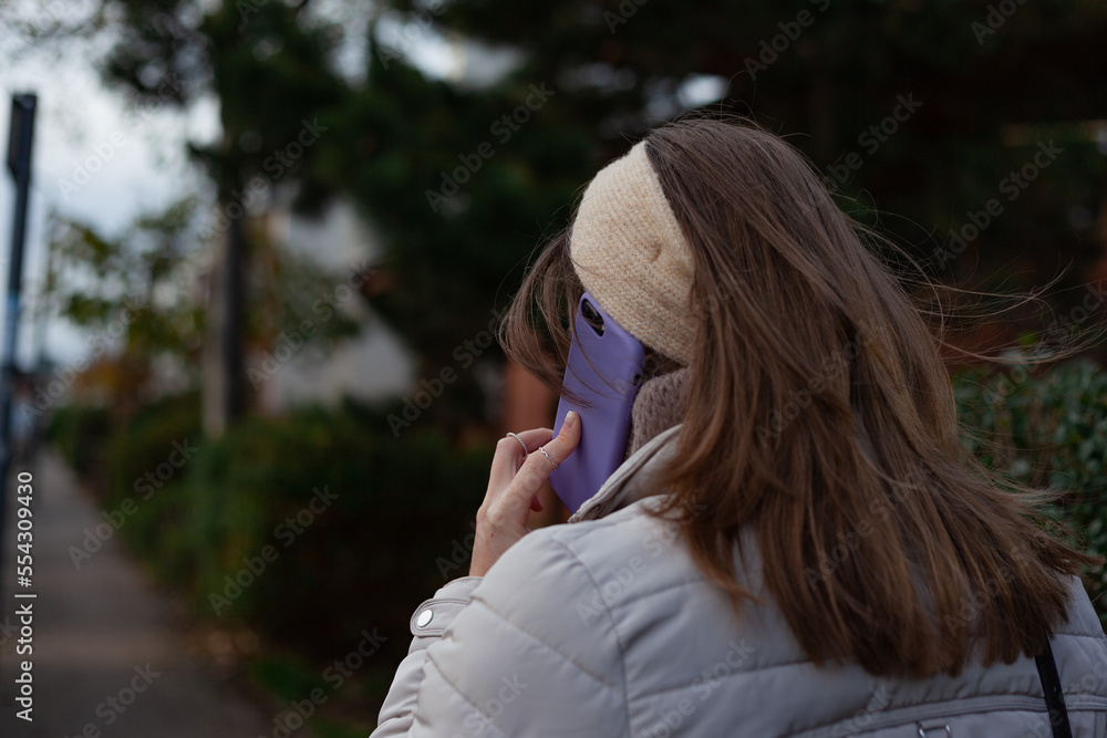 woman talking on cell phone Young woman with long brown hair, wearing a beige jacket and headband, facing opposite direction is walking down the street, speaking her lilac mobile phone and trees 