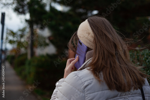 woman talking on cell phone Young woman with long brown hair, wearing a beige jacket and headband, facing opposite direction is walking down the street, speaking her lilac mobile phone and trees 