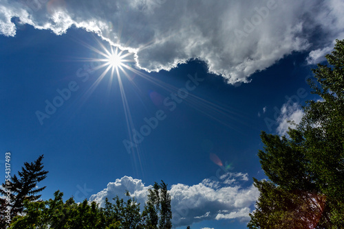 Sun starburst with storm clouds, blue sky and framed by trees in the foreground; Calgary, Alberta, Canada