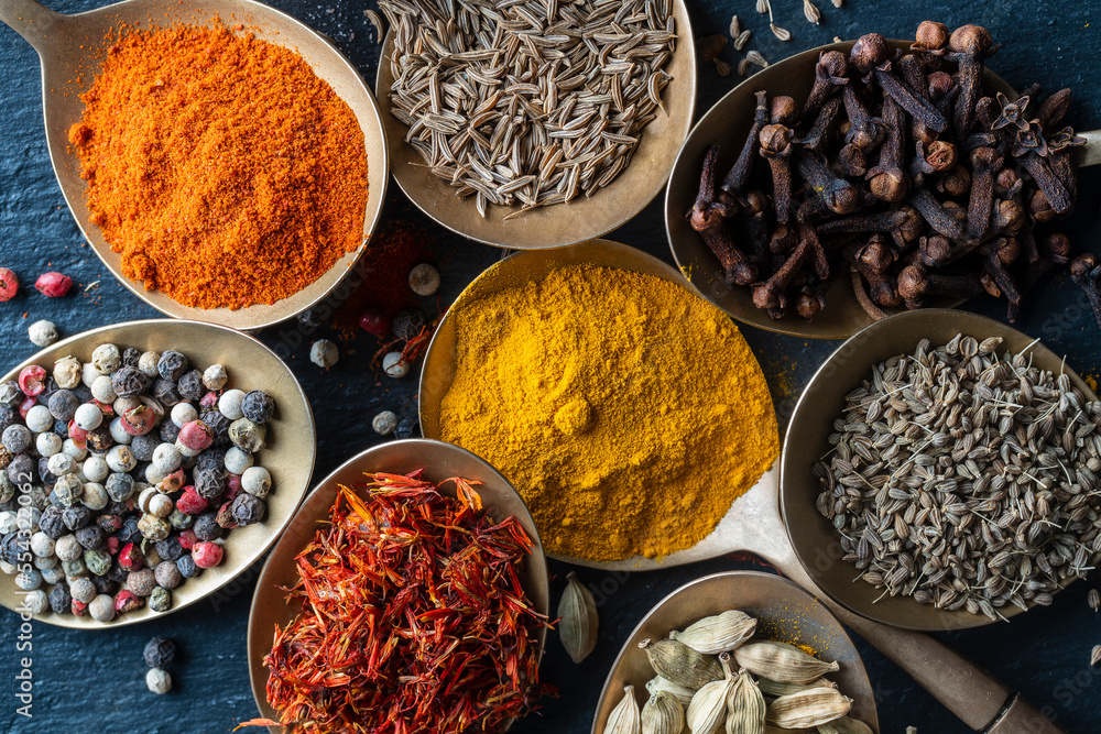 Fototapeta premium Set of spices in brass spoons, closeup, top view. Various peppers, cardamom, carnation, turmeric, paprika and saffron for cooking