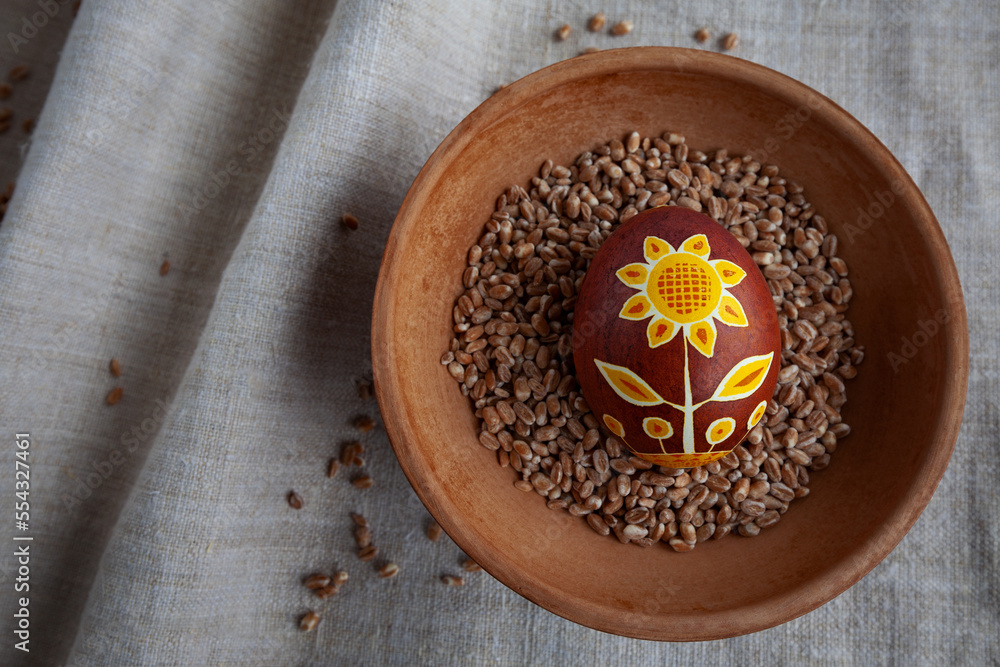 Ukrainian Easter egg in brown-yellow colors with a sunflower flower on a background of linen fabric in a plate with grain Ukrainian Easter egg in brown-yellow colors with a sunflower flower on a background of linen fabric in a plate with grain