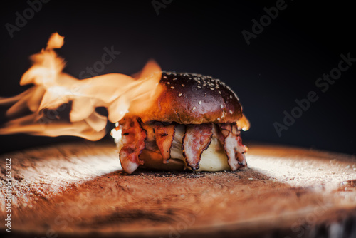  Tasty beef, cheese, burger with many slices of bacon, warm coming out of smoke on a wooden cutting board on a black background, with fire on the stage. 