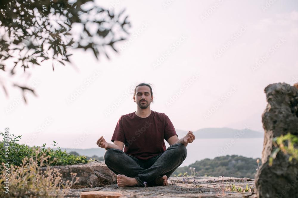 A young man peacefully meditates at sunset in a garden overlooking the mountains and the sea.
