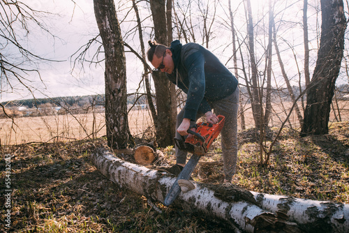 Preparation for the heating season, firewood harvesting. A young man with a chainsaw sawing a fallen tree in the forest.