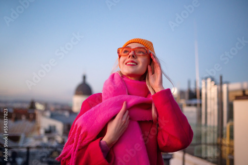 Happy smiling fashionable woman wearing trendy outfit with orange knitted beanie hat, sunglasses, pink woolen fringed scarf, coat, posing outdoor. Copy, empty space for text 