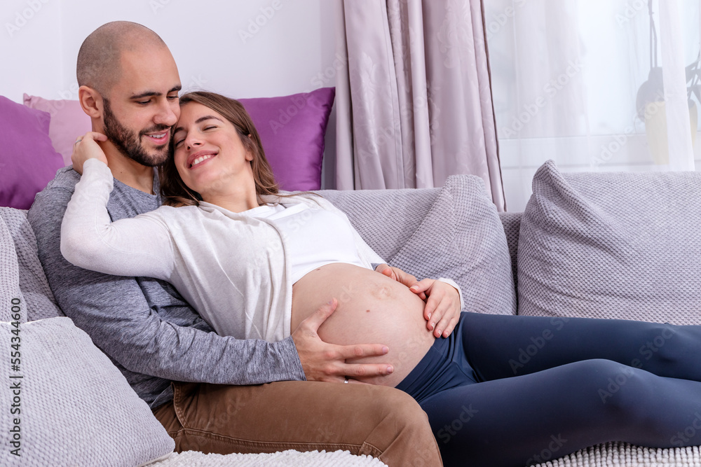 A happy loving couple resting on the sofa, preparing for childbirth. Pregnant woman embraced around her big tummy by her partner. A happy loving couple resting on the sofa, preparing for childbirth. Pregnant woman embraced around her big tummy by her partner.