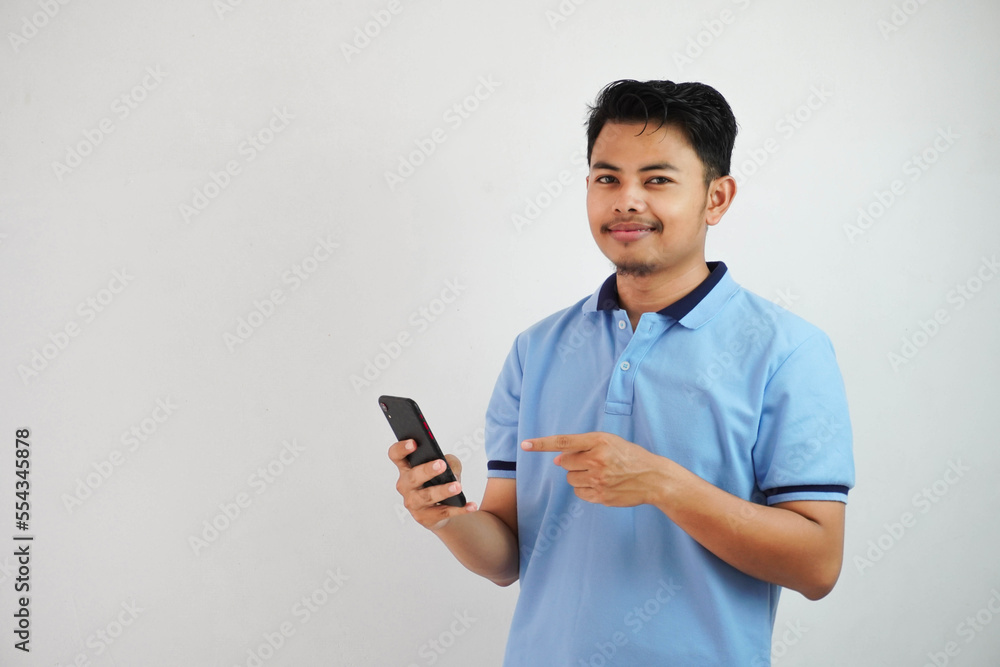 smiling or happy young asian man wearing blue t shirt holding phone and pointing at smartphone with finger, isolated on white background smiling or happy young asian man wearing blue t shirt holding phone and pointing at smartphone with finger, isolated on white background