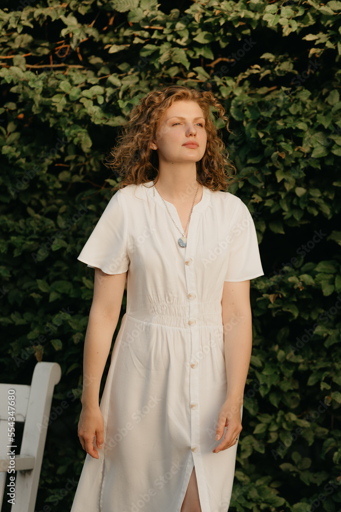 A woman with curly hair is posing in the garden in the evening.