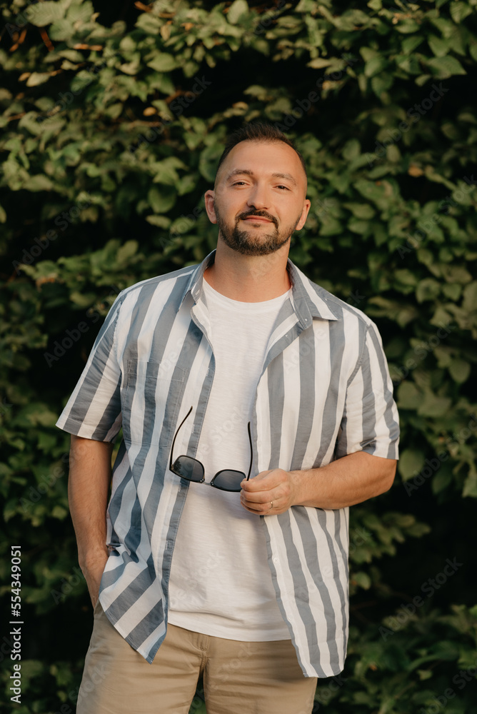 A man is posing in the garden. A guy with green leaves in the background.