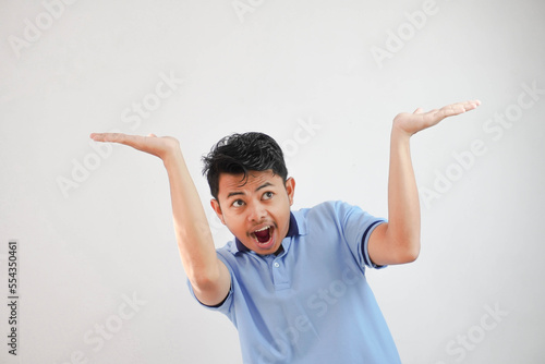 young asian man acting like he's holding up something heavy, but it's just empty copy space wearing blue t shirt isolated on white background. shocked face