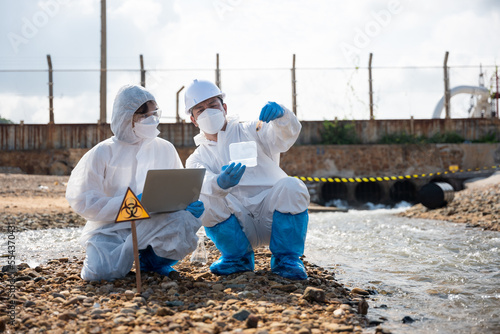 Ecologist sampling taken dead fish to inspaction and save data to laptop computer, Biologist wear protective suit and mask collects sample of waste water from industry, problem environment
