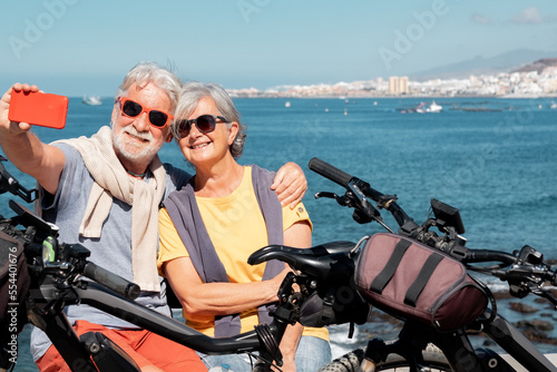 Beautiful happy senior couple in vacation or retirement at sea enjoying healthy lifestyle with bicycles. Sitting outdoors getting a selfie with smartphone