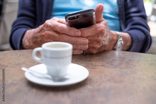Closeup on old senior man hands using mobile phone. Elderly caucasian male sitting at cafe table with an espresso coffee cup while looking at his smartphone