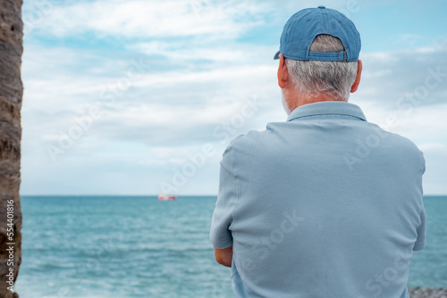Back view of relaxed senior man standing at the beach looking at the horizon over water enjoying free time vacation or retirement. #554401816