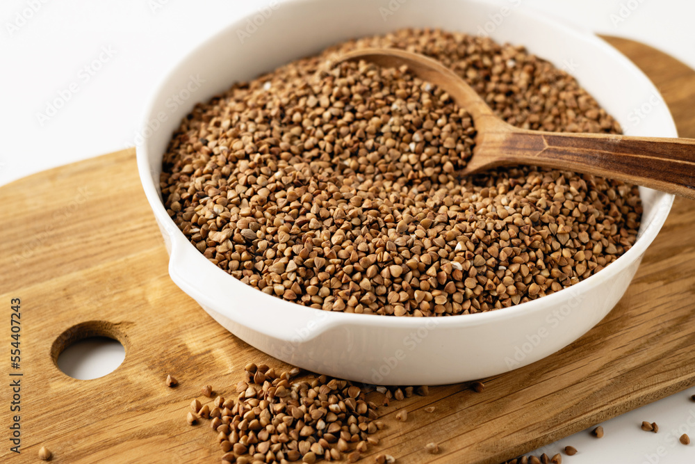 Dry uncooked buckwheat groats in a white dish, wooden board, spoon, on a white background, close-up Dry uncooked buckwheat groats in a white dish, wooden board, spoon, on a white background, close-up