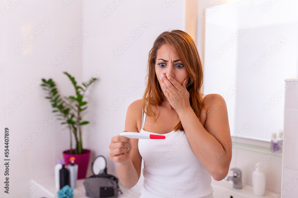 Shocked woman looking at control line on pregnancy test. Shocked young girl with unwanted pregnancy looking the test in the bathroom. sad young woman holding pregnancy test feeling hopeless Shocked woman looking at control line on pregnancy test. Shocked young girl with unwanted pregnancy looking the test in the bathroom. sad young woman holding pregnancy test feeling hopeless