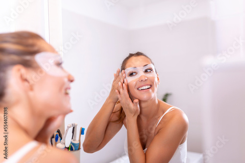 Young beautiful girl applying facial mask on skin. Looking in the mirror in bathroom, Wrapped in a towel, having fun.