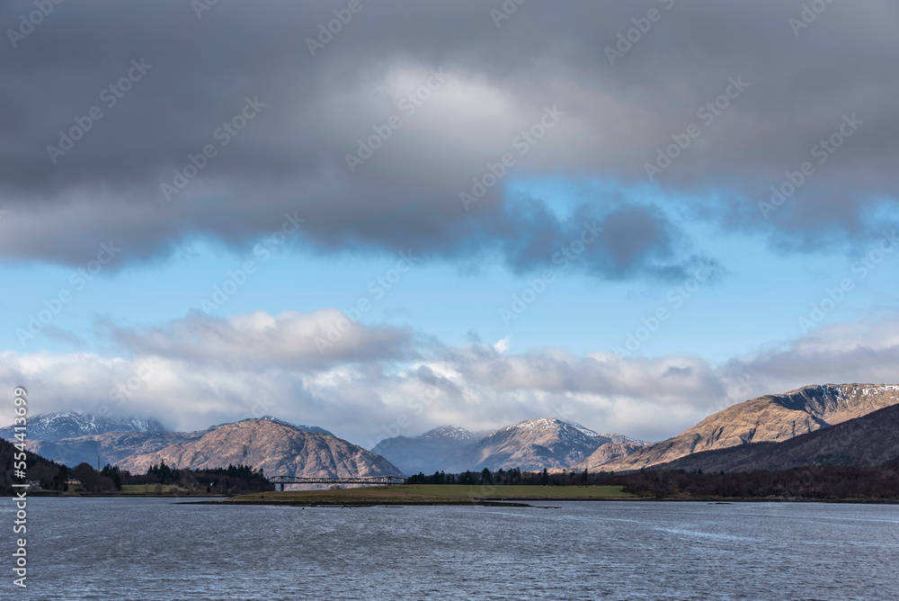 Beautiful Winter landscape view along Loch Leven in Scotland towards snowcapped mountains in distance with dramatic sky