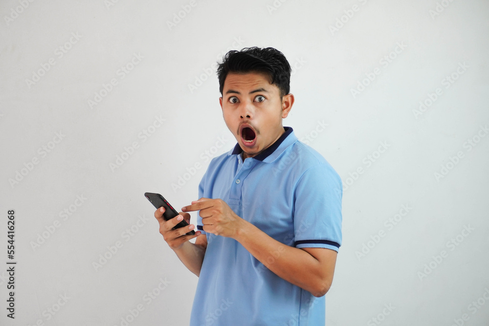 portrait asian man shocked holding phone and pointing at the phone with a finger wearing blue polo t shirt isolated on white background portrait asian man shocked holding phone and pointing at the phone with a finger wearing blue polo t shirt isolated on white background