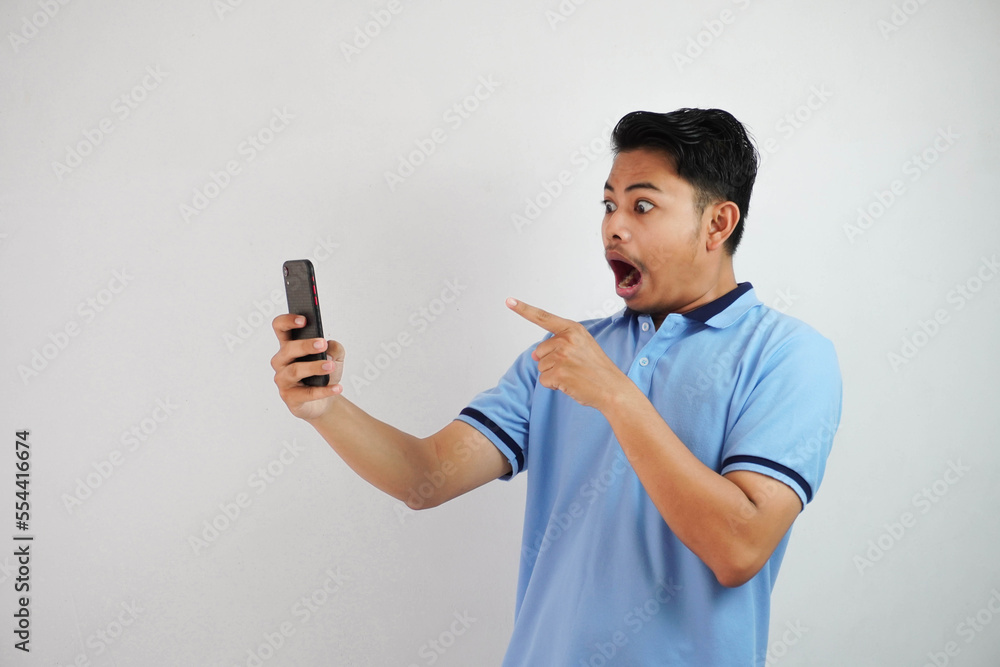 portrait asian man shocked holding phone and pointing at the phone with a finger wearing blue polo t shirt isolated on white background portrait asian man shocked holding phone and pointing at the phone with a finger wearing blue polo t shirt isolated on white background