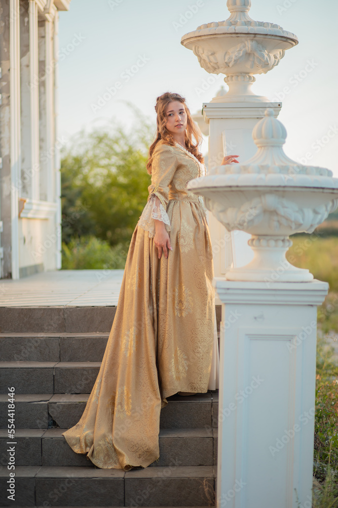Fototapeta premium A beautiful young woman in a historic eighteenth century gold dress stands on the stairs of the mansion. Princess in the palace.